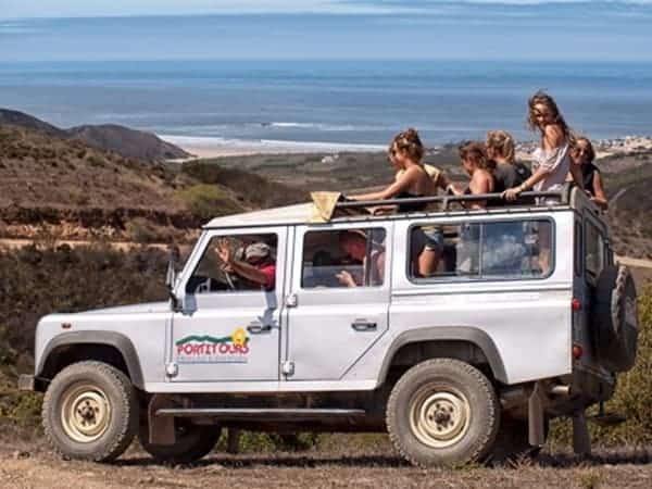 Group in a jeep, on a dirt road with a beach in the back.
